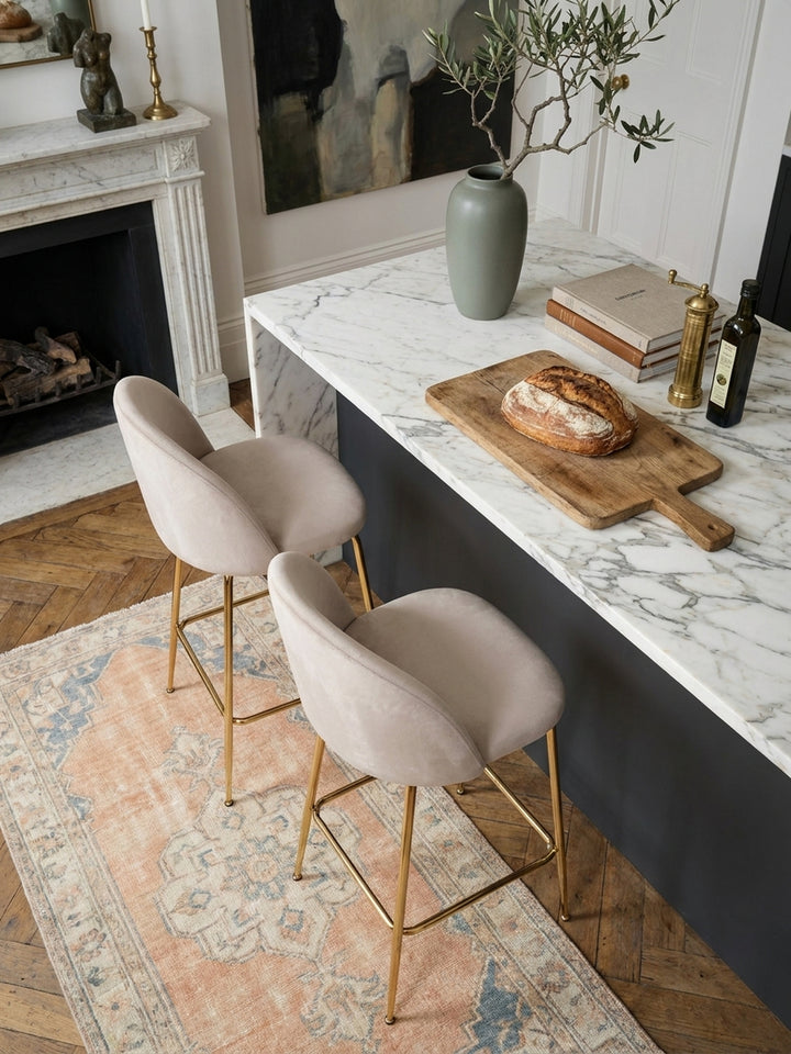 Zwei beige Samt-Küchenhocker mit goldenem Gestell von oben an einer dunklen Marmor-Kücheninsel, arrangiert auf einem Holzboden._