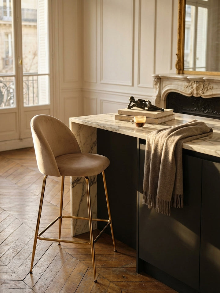 Ein beige Samt-Tresenhocker mit goldenen Beinen neben einem Holztisch, auf dem ein geöffnetes Magazin und eine Kaffeetasse stehen._