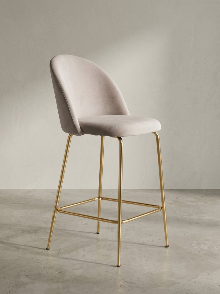Der skandinavische Barhocker in Beige und Gold in einer Dreiviertelansicht, die die geschwungene Rückenlehne und Sitzfläche zeigt._