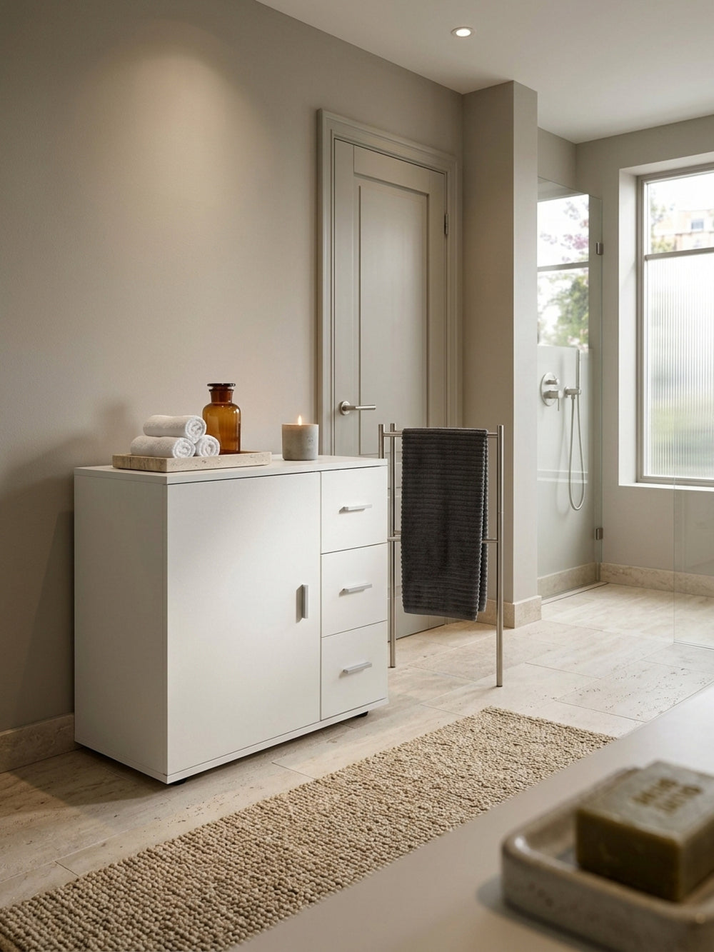 Der weiße Badschrank in einem luxuriösen, warmen Badezimmer mit Travertinboden. Die freistehende 60x66cm Kommode bietet mit Tür und drei Schubladen optimalen Stauraum._