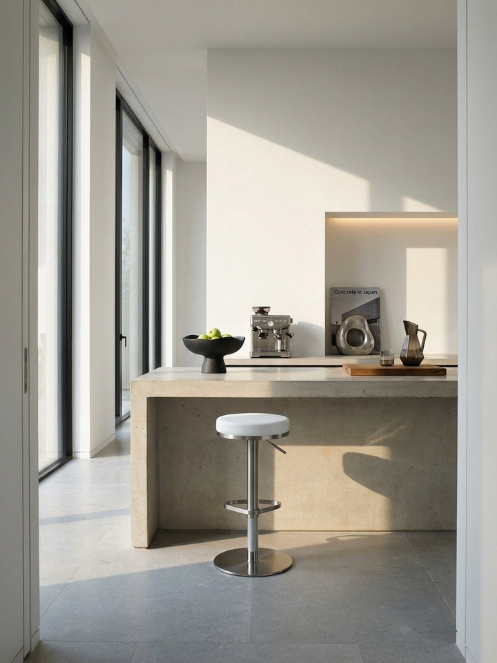 Ein puristischer Barhocker in Weiß mit Edelstahlgestell steht an einer modernen Kücheninsel aus grauem Beton im lichtdurchfluteten Penthouse._