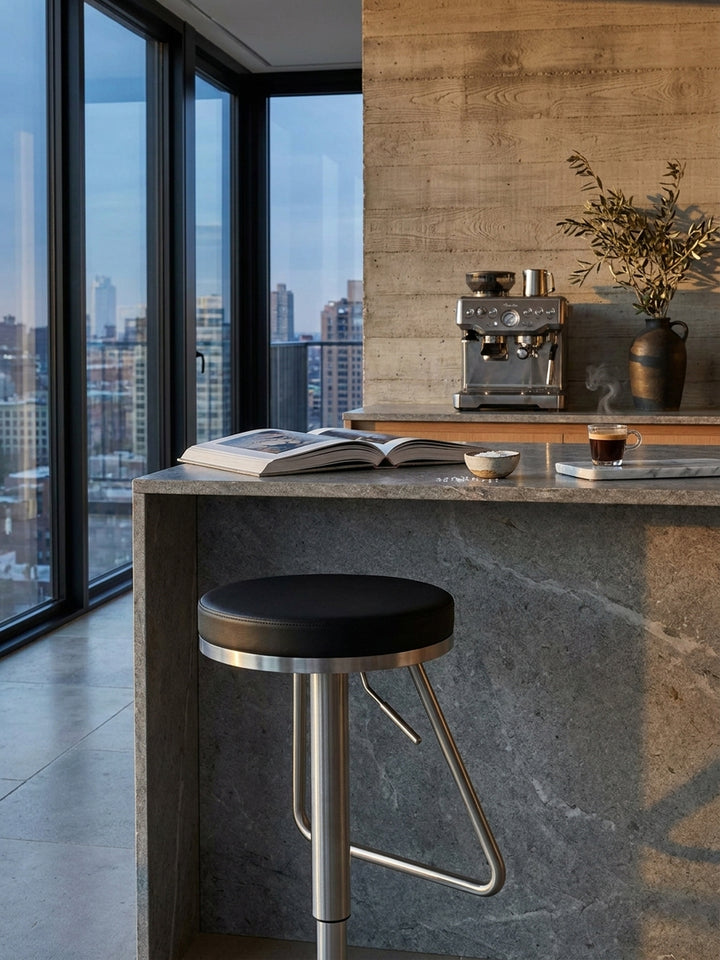 Höhenverstellbarer Bistrohocker platziert an einer glatten Natursteinplatte neben einem aufgeschlagenen Architektur-Bildband und einer Espressotasse._