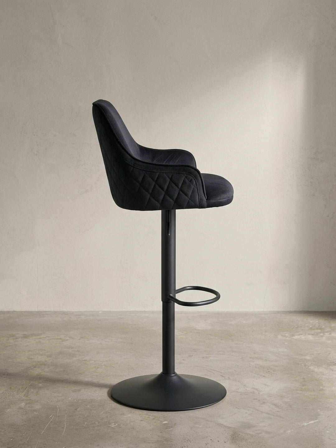 Seitenansicht des schwarzen Samt-Hockers, die das schlanke Profil, die Höhe der Rückenlehne und die ergonomische Position der integrierten Fußstütze verdeutlicht._