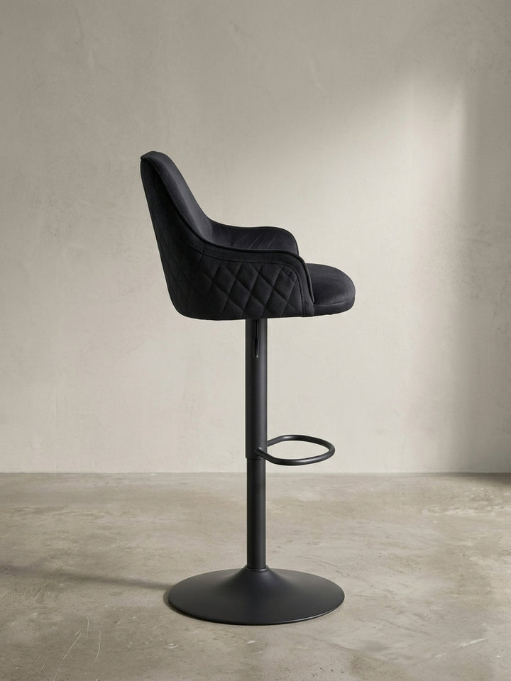 Seitenansicht des schwarzen Samt-Hockers, die das schlanke Profil, die Höhe der Rückenlehne und die ergonomische Position der integrierten Fußstütze verdeutlicht._