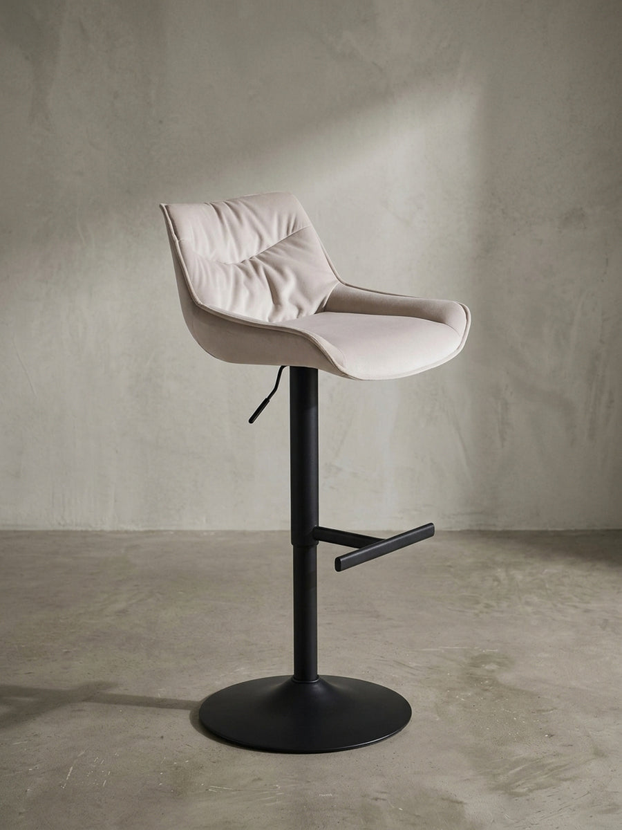 Moderner Design-Barhocker in 3/4-Ansicht, der die geschwungene Sitzschale aus beigem Samt und die praktische Fußstütze aus schwarzem Metall zeigt._