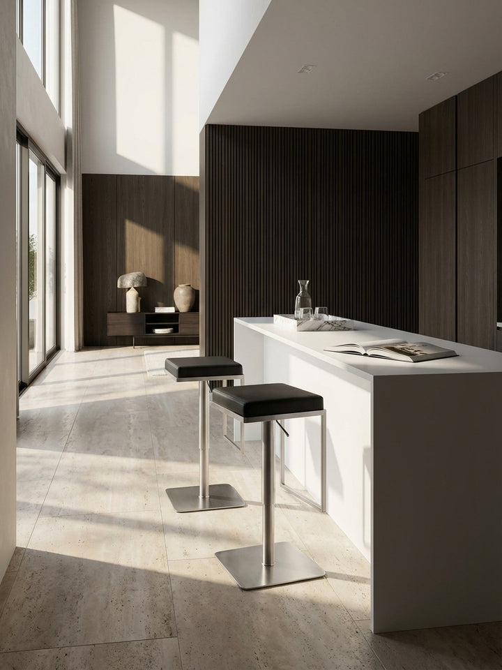 Moderner Barhocker aus schwarzem Kunstleder und Edelstahl an einer minimalistischen Kücheninsel aus weißem Corian vor einer dunklen Eichenholzwand._