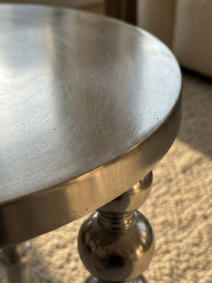 Nahaufnahme der spiegelnden Oberfläche des silbernen Metall-Beistelltisches, die das Licht einfängt und die hochglanzpolierte Textur des Aluminiums zeigt._