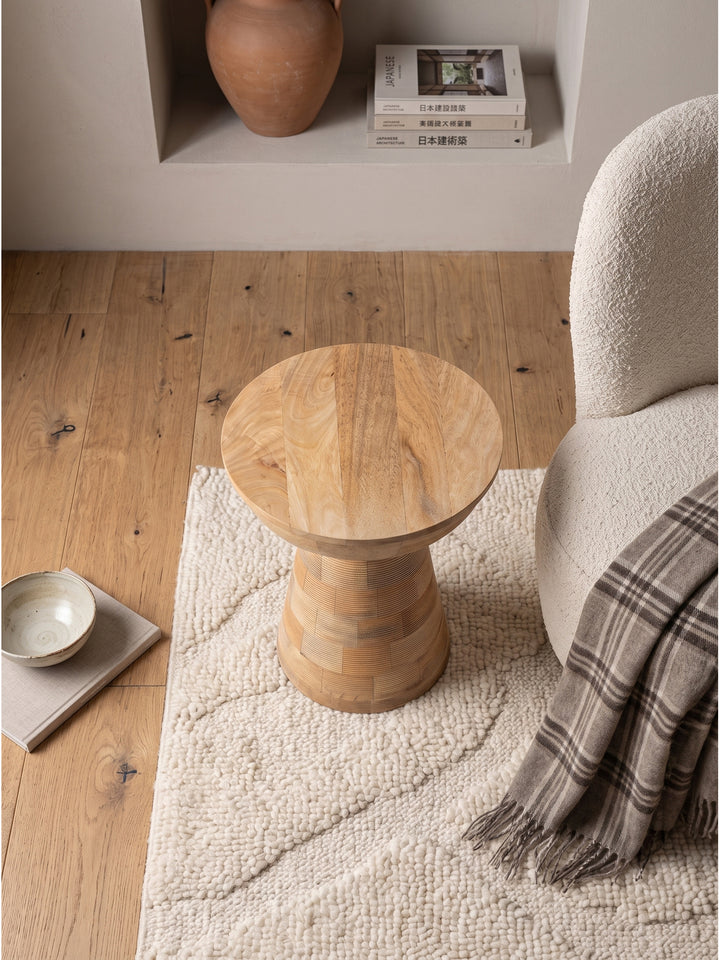 Draufsicht auf den runden Holztisch aus natürlichem Mango Massivholz im modernen Wohnbereich, arrangiert mit einer skulpturalen Stehlampe und einem hellen Sessel auf edlem Eichenparkett_