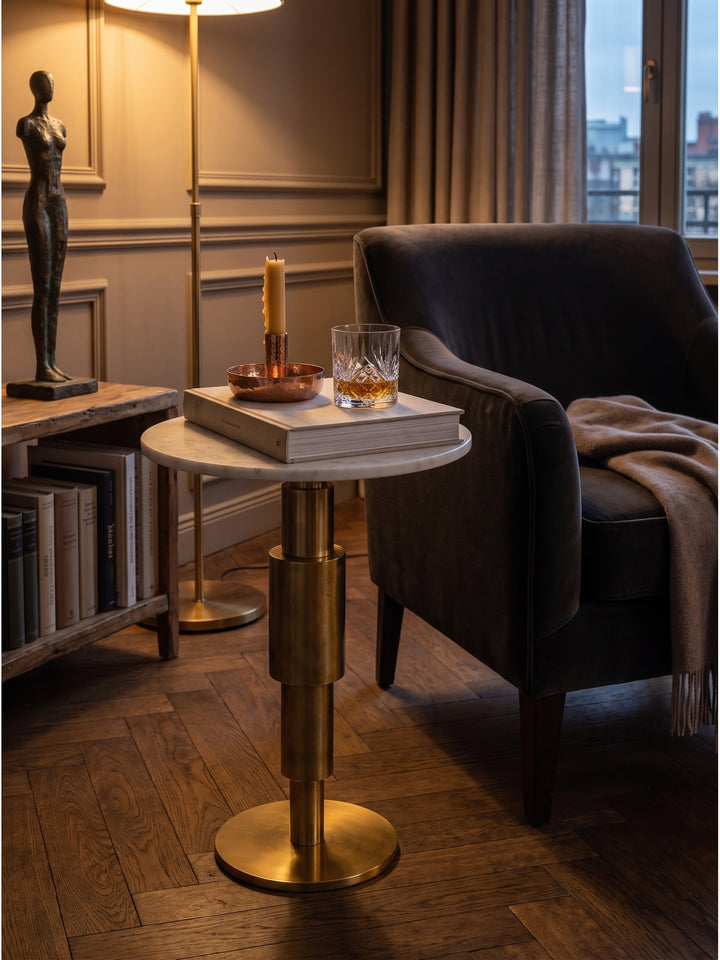 Goldener Anstelltisch aus texturiertem Aluminium dekoriert mit einem schweren Kristallglas neben einem dunklen Lesesessel im eleganten Wohnraum_