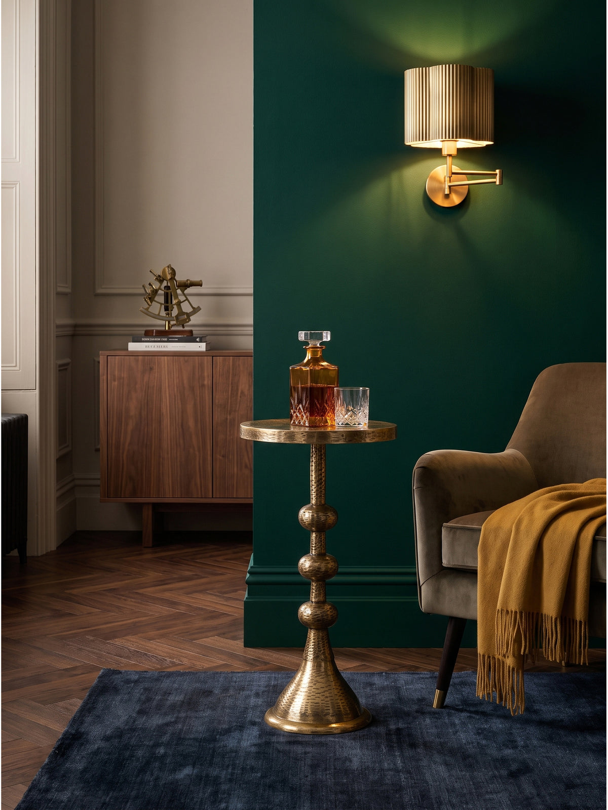 Ein bernsteinfarbener Glaskaraffe und ein Kristallglas stehen auf dem kleinen goldenen Anstelltisch aus Metall direkt an der Armlehne des Wohnzimmersofas._