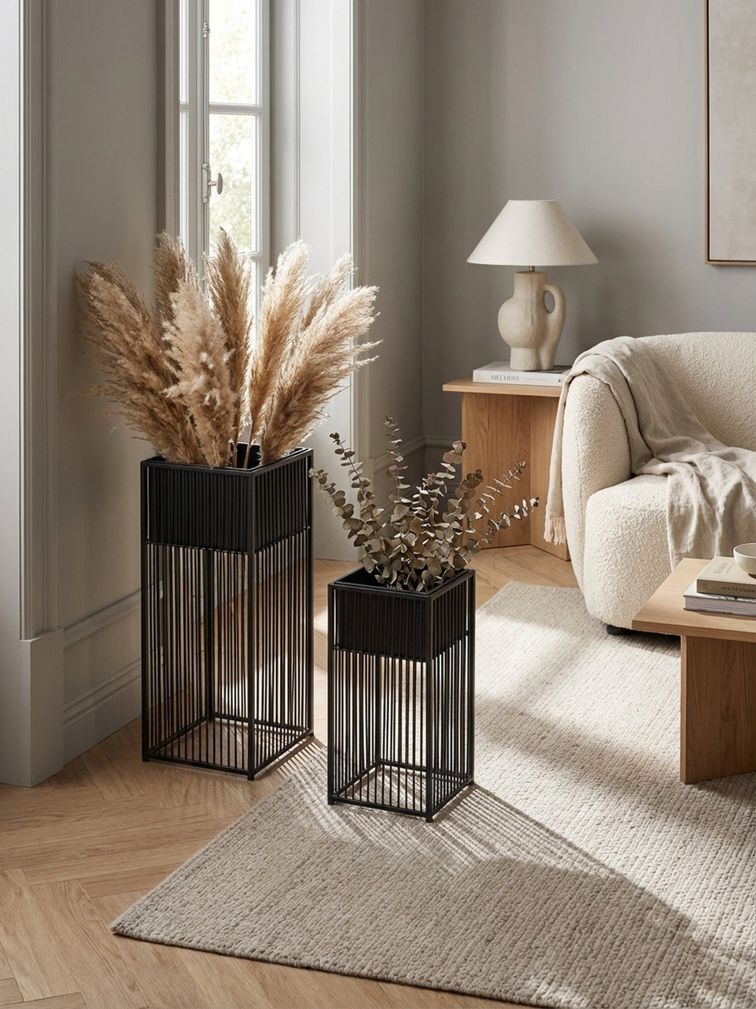 Draufsicht auf ein stilvolles Interieur, das ein 2er Set schwarzer Pflanzgefäße aus Metall auf einem beigen Wollteppich neben einem Sofa und einem Beistelltisch zeigt._