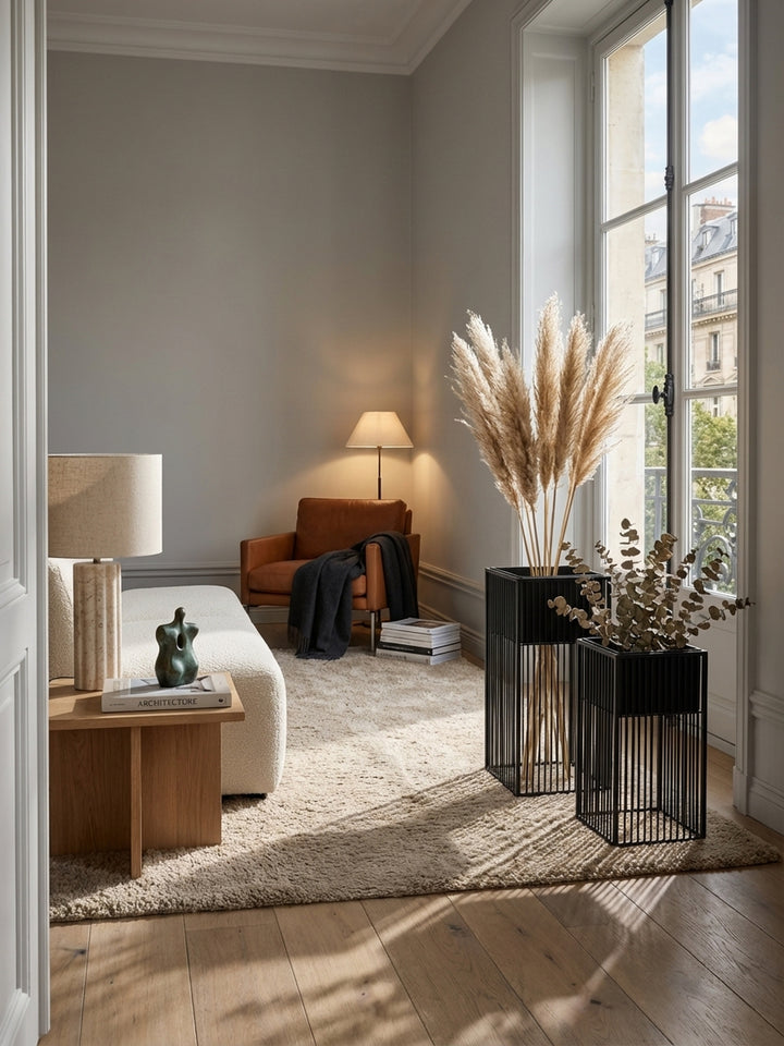 Modernes Wohnzimmer mit zwei schwarzen Metall-Pflanzkübeln neben einem großen Fenster, die mit Trockenblumen dekoriert sind und Schatten auf den Holzboden werfen._