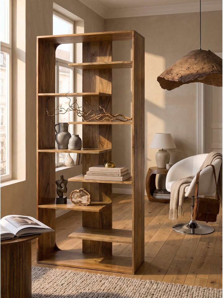 Offenes Bücherregal aus Mangoholz kombiniert mit einem skulpturalen Travertin-Couchtisch und weichem Sessel im sonnigen Wohnzimmer_