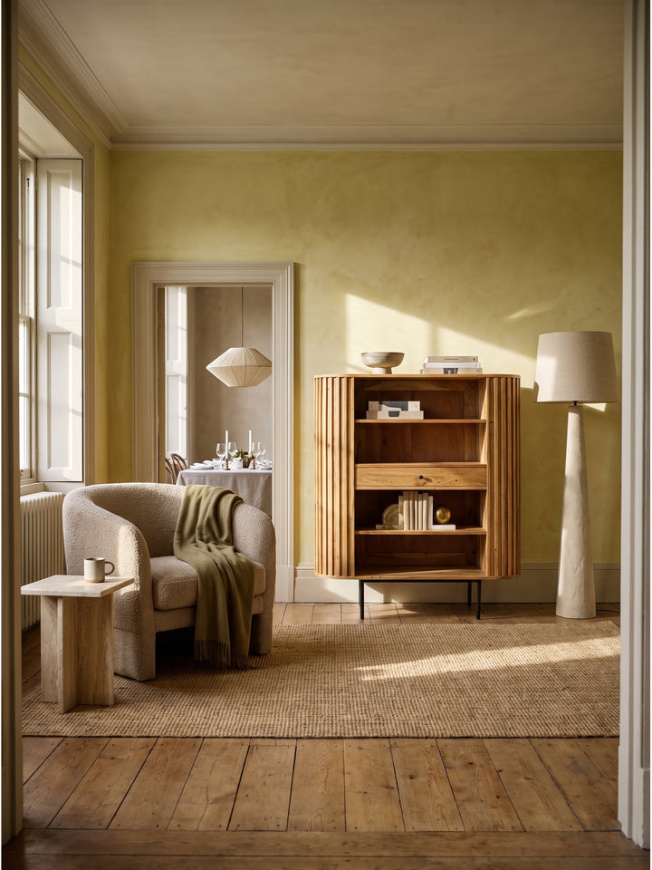 Bücherregal aus massivem Akazienholz mit Lamellen-Design 105x122 cm freistehend im modern eingerichteten Wohnzimmer mit warmweißen Wänden._