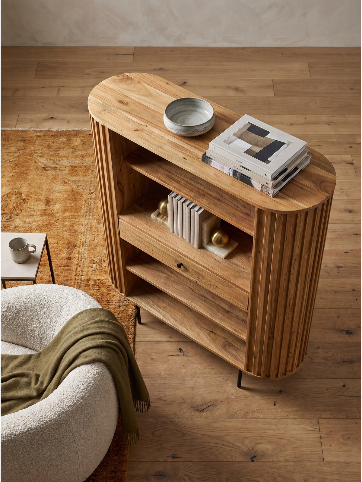 Blick von oben auf das Bücherregal aus massiver Akazie mit runden Ecken im Organic Modern Wohnzimmer neben einem Bouclé-Sessel._