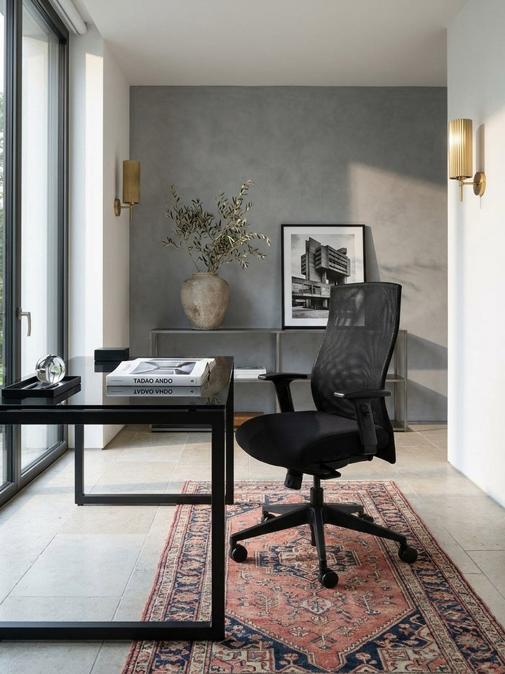 Schwarzer Design Schreibtischstuhl gepaart mit einem dunklen Holzschreibtisch und einem ledernen Loungesessel im Hintergrund des eleganten Arbeitsbereichs._