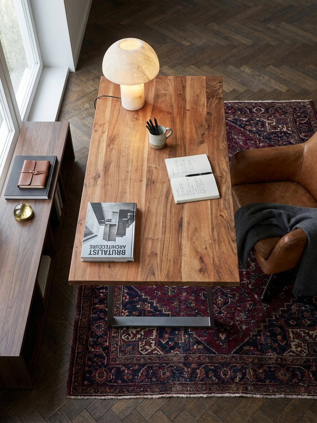 Draufsicht auf einen modernen Bürotisch aus Akazie mit Metallbeinen auf einem Vintage-Perserteppich, arrangiert mit einem Leder-Bürostuhl und einer Messing-Tischlampe._