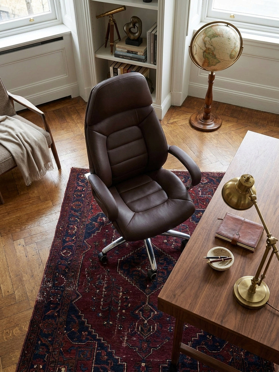 Blick von schräg oben auf einen ergonomischen Drehstuhl mit braunem Lederbezug an einem Holztisch, arrangiert auf einem antiken Perserteppich im Arbeitszimmer._