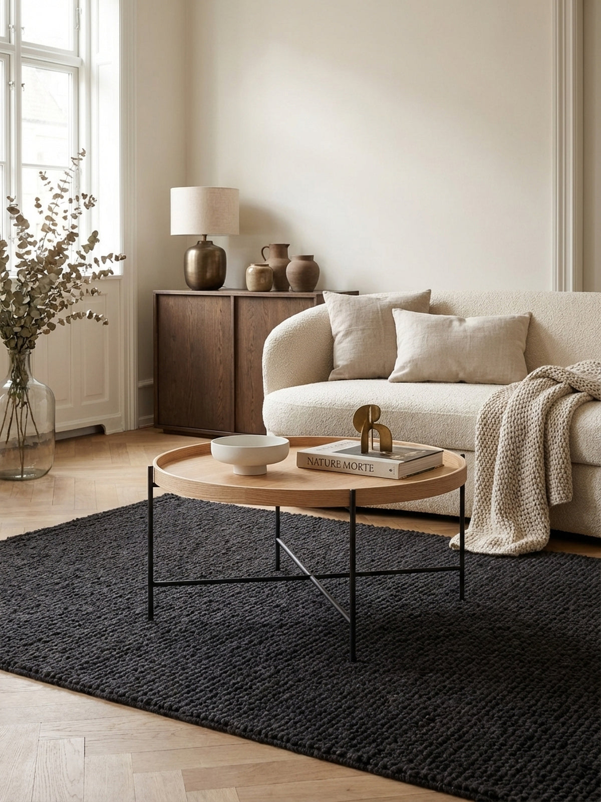 Runder Couchtisch aus Eichenholz mit filigranem Metallgestell als Mittelpunkt einer stilvollen Wohnzimmer-Sitzgruppe auf einem dunklen Teppich vor einem Sofa.