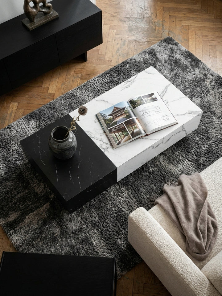 Von oben fotografierter Loungetisch mit asymmetrischem Design, der vor einer großen Wohnlandschaft auf einem texturierten grauen Teppich steht._