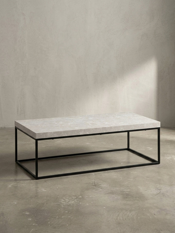 Schräge 3/4-Ansicht des Design-Sofatisches, die die Tiefe der 5 cm starken Tischplatte in Stein-Optik und die filigrane Struktur des Eisengestells betont._