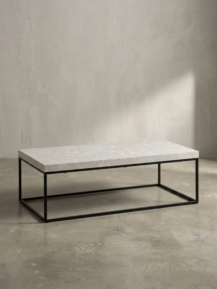 Schräge 3/4-Ansicht des Design-Sofatisches, die die Tiefe der 5 cm starken Tischplatte in Stein-Optik und die filigrane Struktur des Eisengestells betont._
