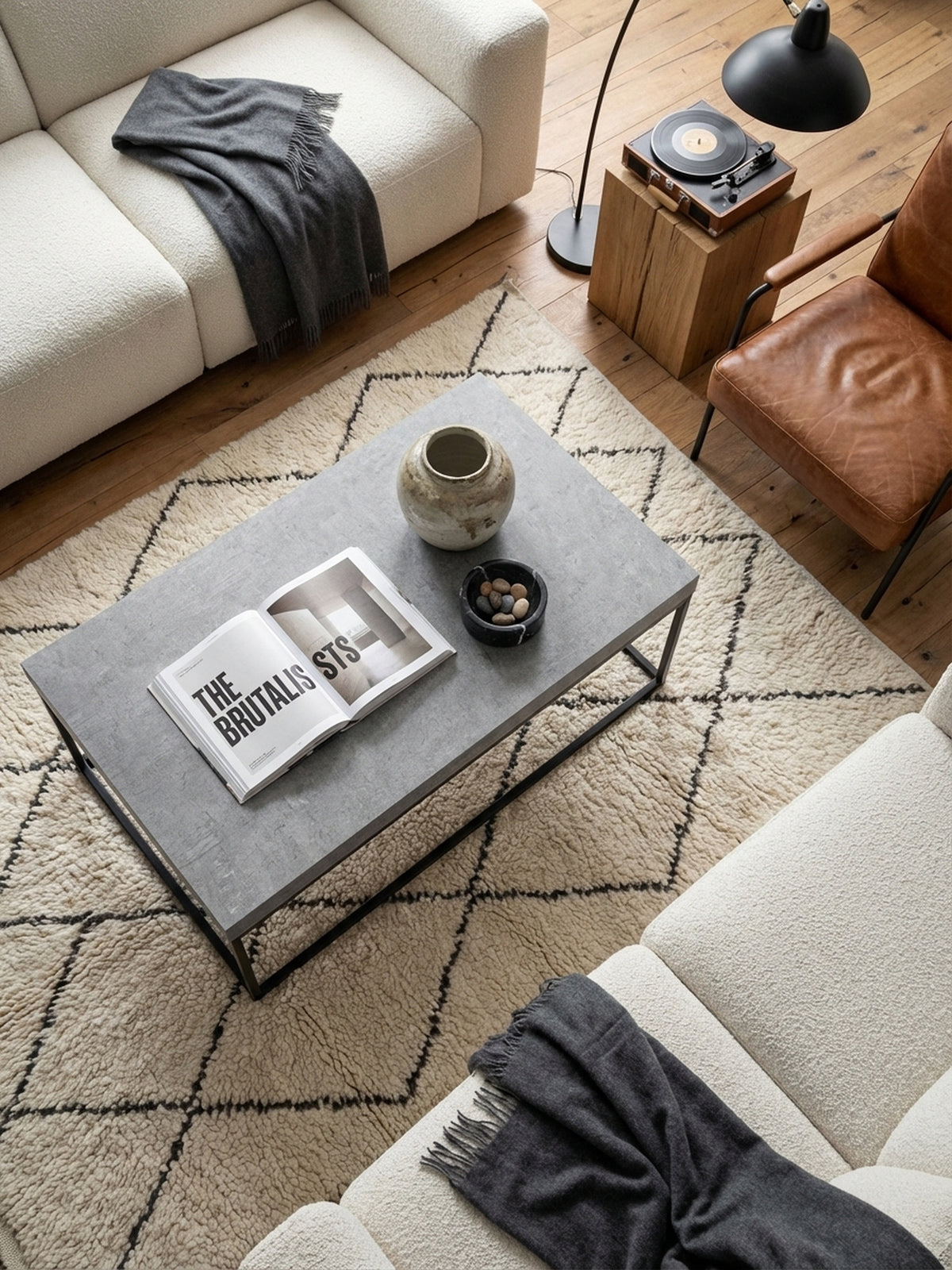 Draufsicht auf einen Loungetisch mit grauer Platte und schwarzem Metallgestell, arrangiert auf einem cremefarbenen Wollteppich vor einem Sofa._