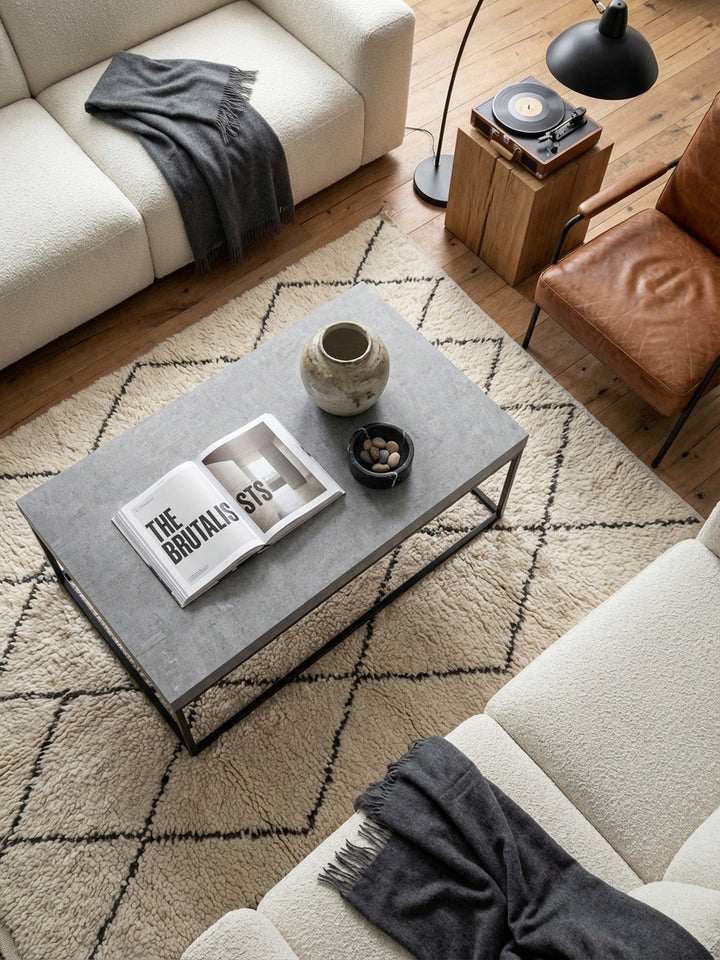 Draufsicht auf einen Loungetisch mit grauer Platte und schwarzem Metallgestell, arrangiert auf einem cremefarbenen Wollteppich vor einem Sofa._