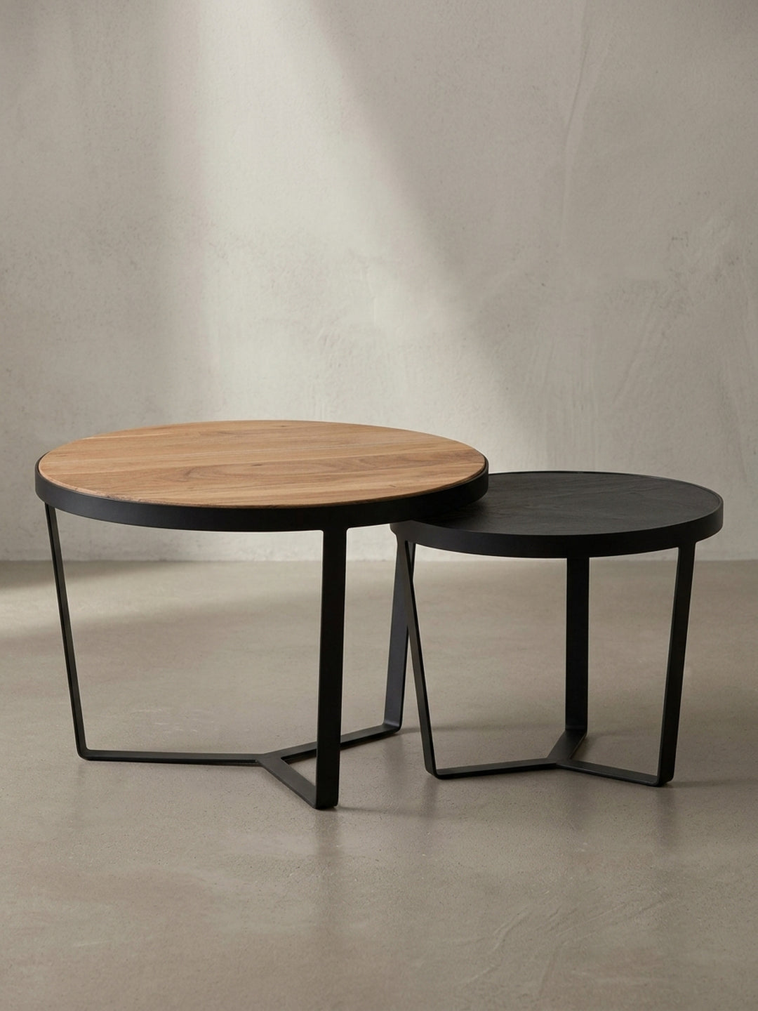 Freigestellter Couchtisch mit runder Akazienholz-Platte und schwarzem Metallgestell, der kleinere Satztisch in Steinoptik ist daneben platziert._