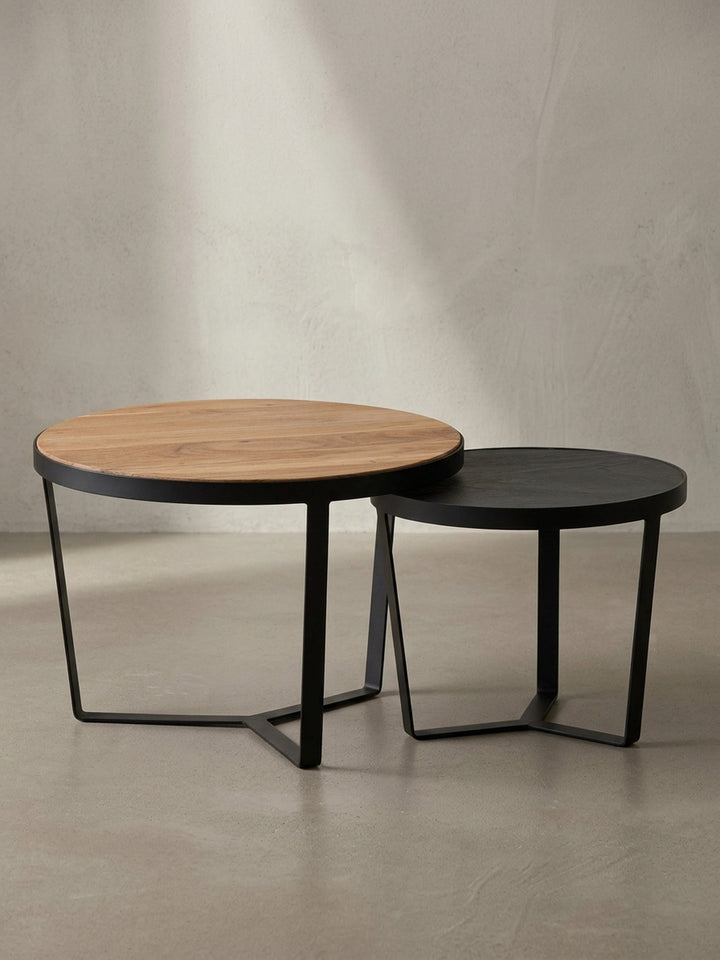 Freigestellter Couchtisch mit runder Akazienholz-Platte und schwarzem Metallgestell, der kleinere Satztisch in Steinoptik ist daneben platziert._
