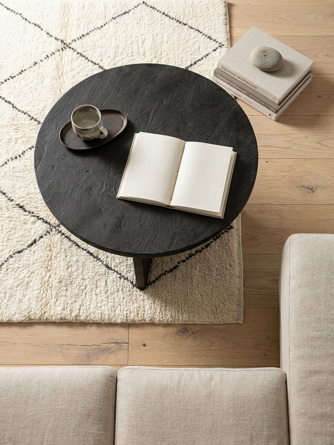 Blick von oben auf den schwarzen Sofatisch mit drei Beinen auf einem Jute-Teppich, arrangiert vor einer minimalistischen Couch in einem organisch-modernen Ambiente._