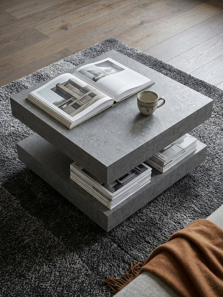 Quadratischer Sofatisch in Beton-Optik von oben gesehen, arrangiert auf einem beigen Teppich vor einer großen Wohnlandschaft in einem hellen Raum._