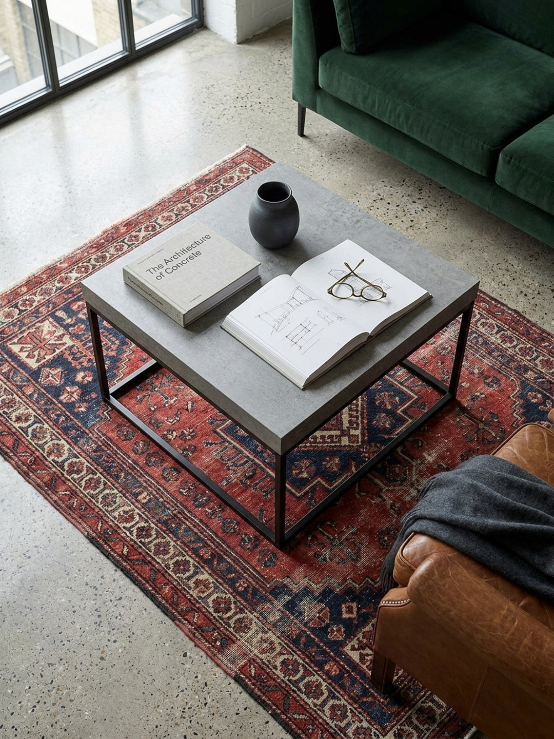 Quadratischer Sofatisch mit grauem Betondekor von oben gesehen, arrangiert mit einer Vase und einem Buch vor einer modernen Wohnlandschaft._