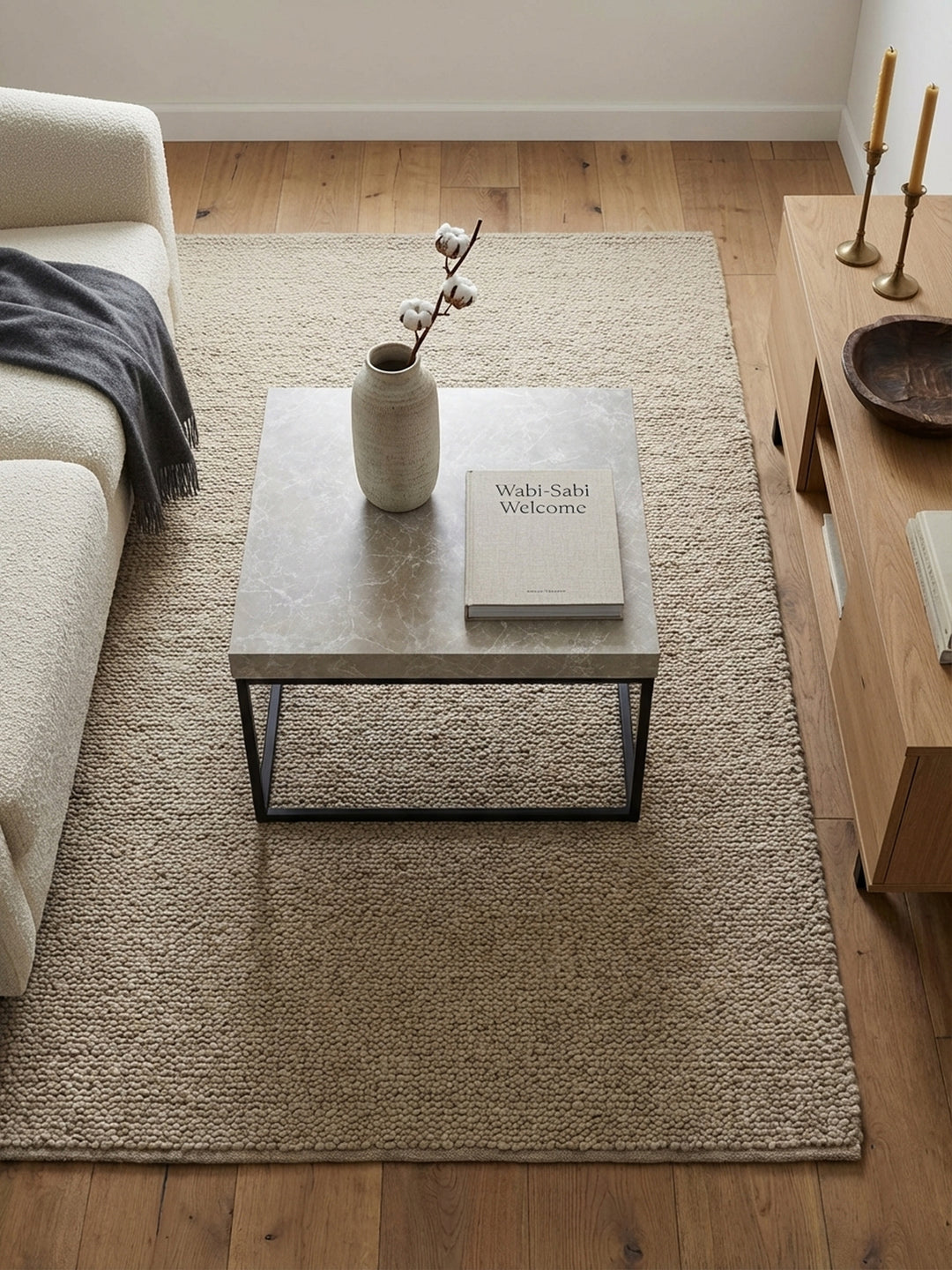 Quadratischer Sofatisch mit schwarzem Metallgestell von oben betrachtet auf einem sandfarbenen Wollteppich, arrangiert vor einer minimalistischen Couch._