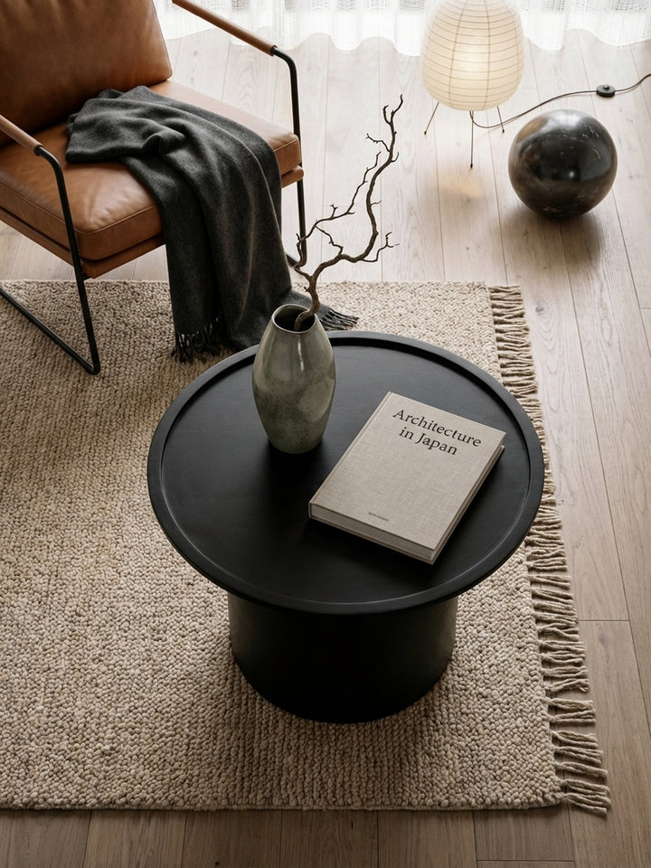 Runder Sofatisch aus schwarzem Massivholz von oben, arrangiert auf einem cremefarbenen Teppich vor einer modernen Wohnlandschaft._