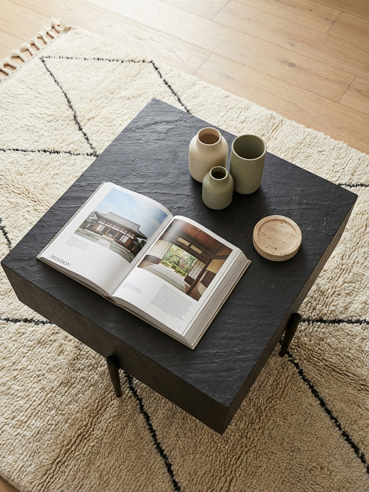 Von oben gesehen, der anthrazitfarbene Design-Couchtisch auf einem flauschigen Wollteppich, perfekt arrangiert vor einem minimalistischen Sofa aus Leinenstoff.