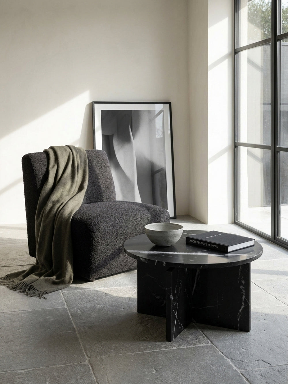 Geometrischer Wohnzimmertisch mit Kreuzgestell arrangiert neben einer modernen puristischen Couch und einer skulpturalen Travertin-Stehlampe_