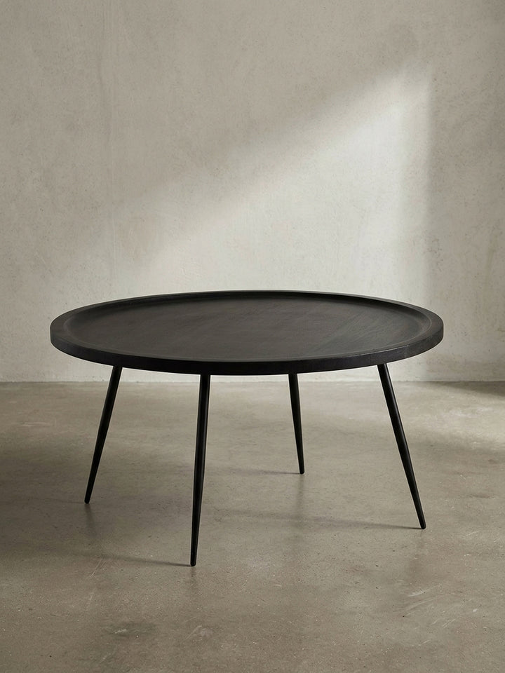 Freigestellte Frontansicht des runden Couchtischs aus massivem Mangoholz in Schwarz mit vier filigranen Metallbeinen vor weißem Hintergrund._