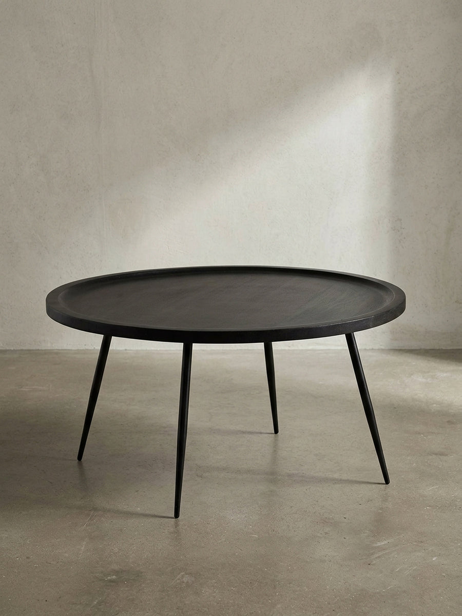 Freigestellte Frontansicht des runden Couchtischs aus massivem Mangoholz in Schwarz mit vier filigranen Metallbeinen vor weißem Hintergrund._