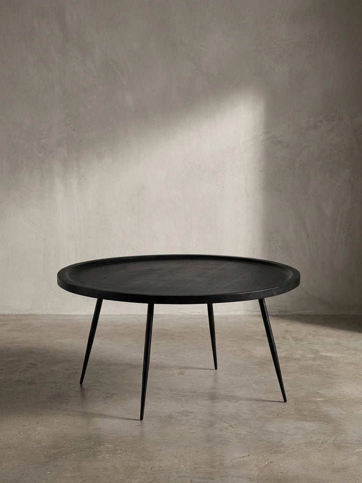 Schräger Blick auf den zeitlosen Sofatisch mit runder Platte aus schwarzem Mangoholz, der seine elegante Form und das schlanke Beingestell betont._