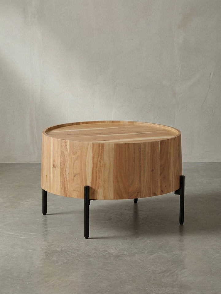 Frontalansicht des runden Couchtisches aus massivem Akazienholz mit seinem charakteristischen Trommelkorpus und den filigranen, schwarzen Metallbeinen vor weißem Hintergrund._