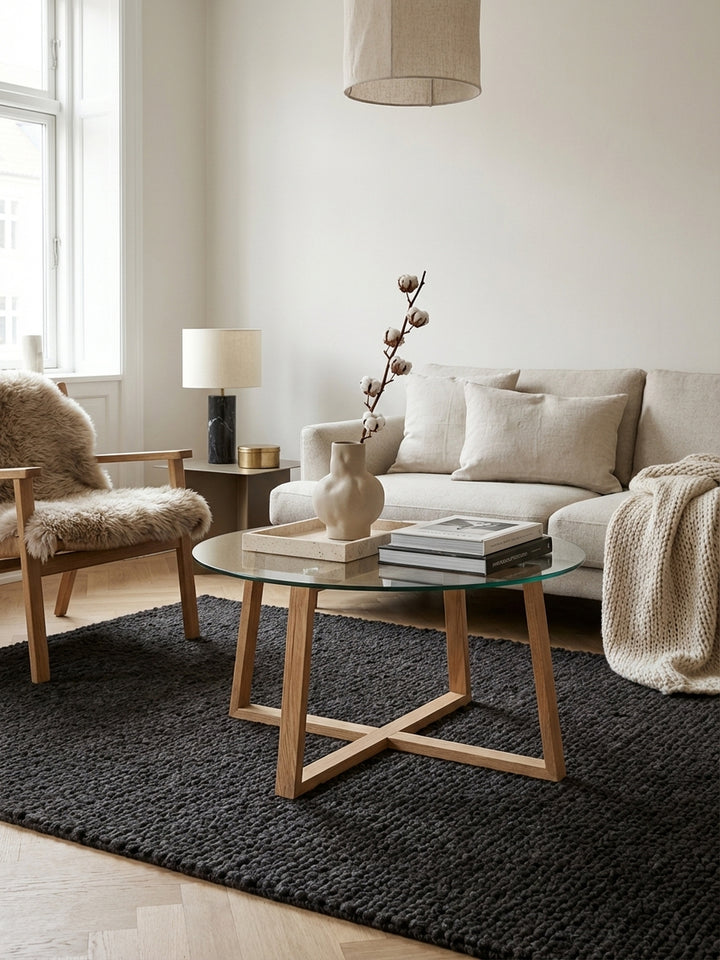 Runder Couchtisch mit Glasplatte und massivem Eichenholzgestell in einem modern eingerichteten, hellen Wohnzimmer, dekoriert mit Büchern und Tassen._