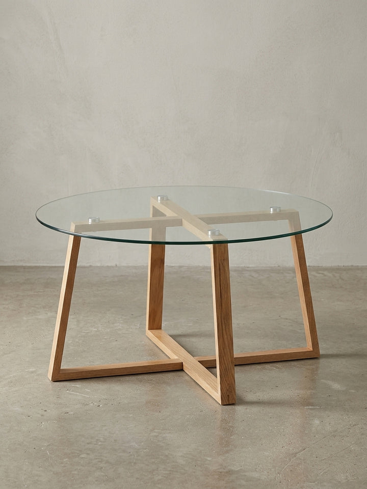 Moderner runder Couchtisch mit einem Gestell aus massivem Eichenholz und einer Glasplatte, freigestellt vor weißem Hintergrund in Frontalansicht._