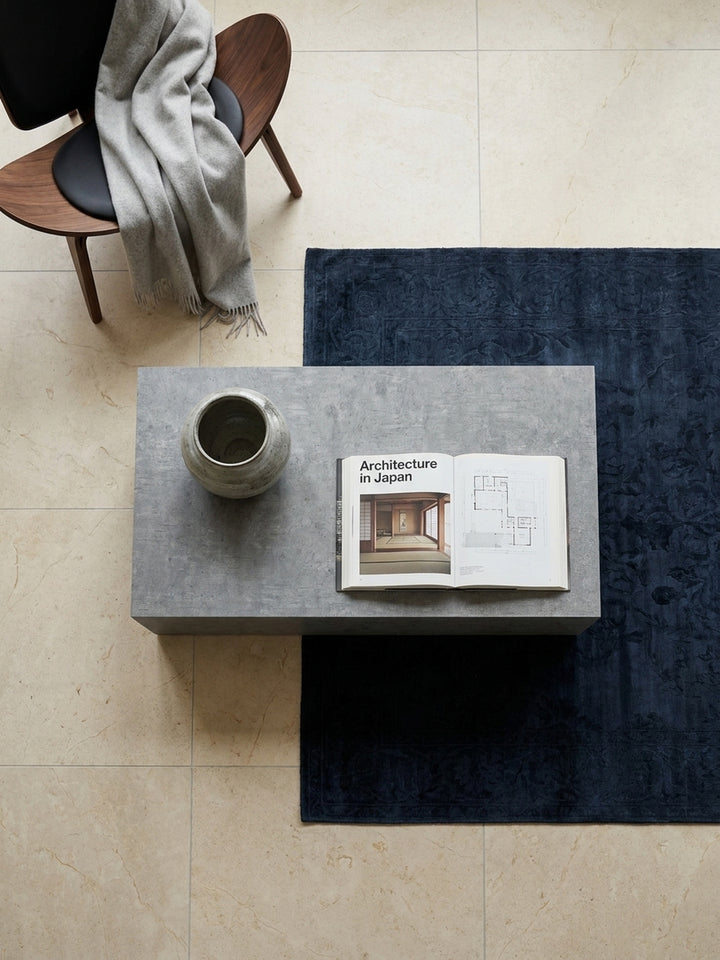 Draufsicht auf einen rechteckigen Wohnzimmertisch im Beton-Design, der symmetrisch auf einem silbergrauen Seidenteppich platziert ist._