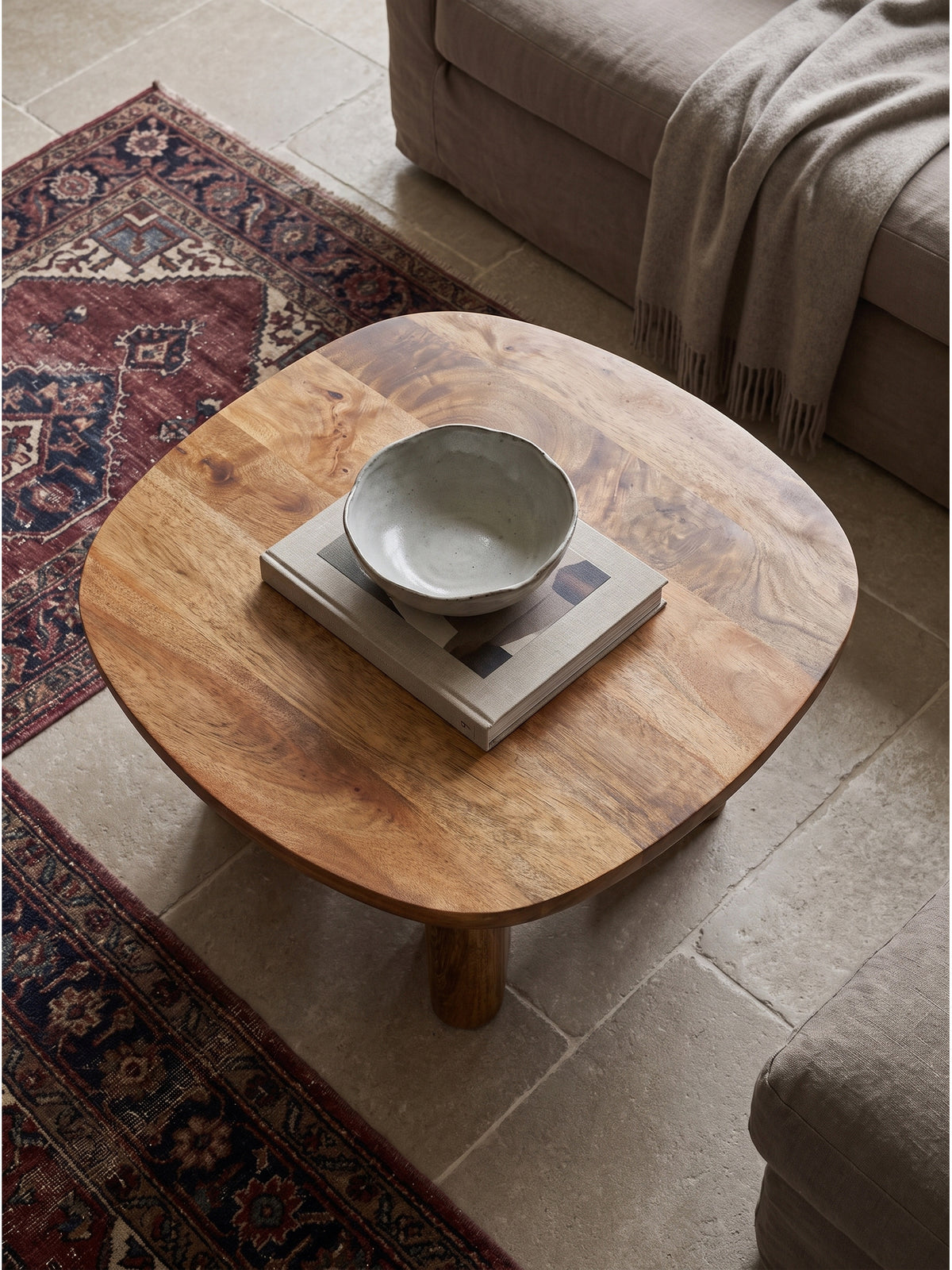 Draufsicht auf den quadratischen Massivholz Couchtisch neben einem hellen Leinensofa mit drapiertem Kaschmirplaid auf edlen warmen Kalksteinfliesen.