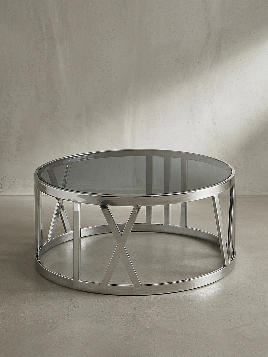 Silberner Couchtisch mit einer runden Platte aus schwarz getöntem Glas und einem geometrischen Edelstahlgestell freigestellt vor weißem Hintergrund._