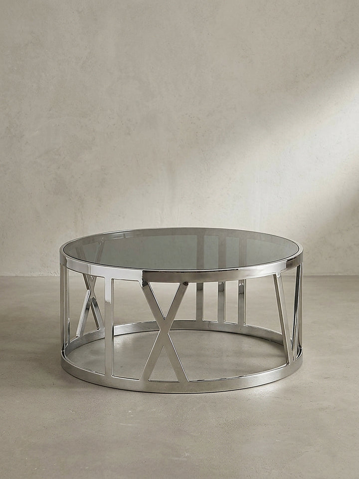 Runder Couchtisch aus Metall und schwarzem Glas im neutralen Studiolicht, das die symmetrische und offene Konstruktion des Gestells klar hervorhebt._