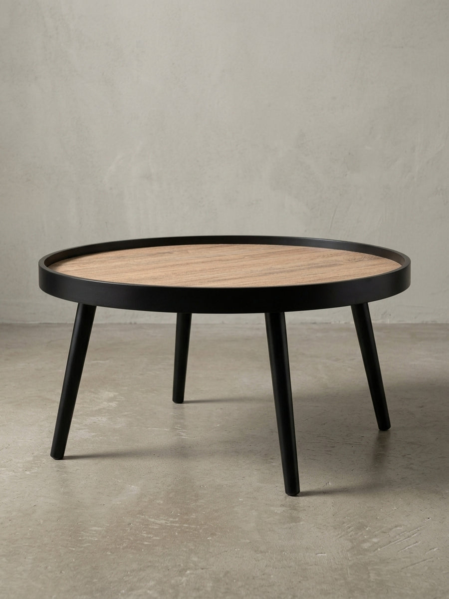 Schräger Blick auf den runden Salontisch, der die erhöhte Kante der Tischplatte und die elegant gespreizten schwarzen Holzbeine zeigt._