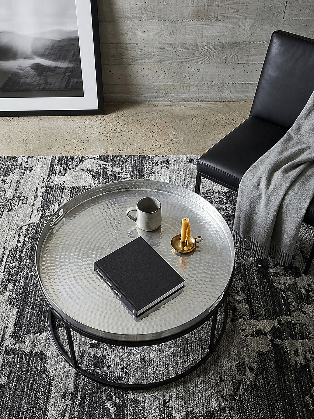 Draufsicht auf den silbernen Tabletttisch aus Metall neben einem schwarzen Ledersessel in einer minimalistischen Lounge mit kühlen grauen Wänden._