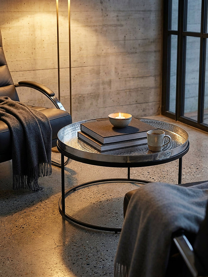 Silberner Sofatisch aus Metall mit abnehmbarem Tablett dekoriert mit einer Keramiktasse und Bildband vor einer dunklen Betonwand._